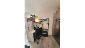 Un dormitorio pequeño con cama, escritorio y silla. Se observa un armario con espejo y una estantería con una planta en maceta.