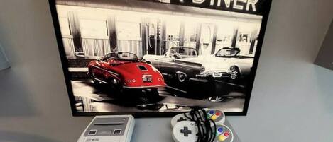 Un téléviseur à écran plat posé sur un meuble, avec une affiche sur le thème de l’automobile, une console de jeux et des manettes.