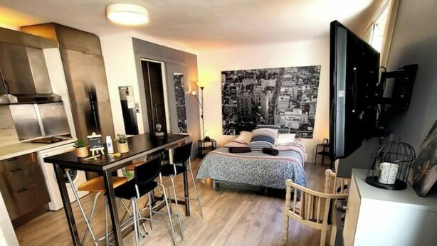 Habitación de hotel compacta con kitchenette, cama con un mural de paisaje urbano, televisión y un pequeño rincón de estar.