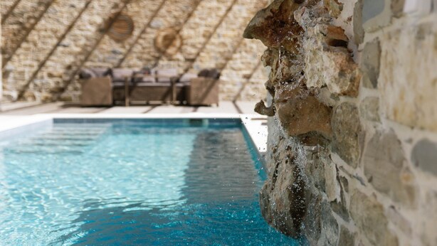 Une piscine à l’eau d’un bleu cristallin, entourée de murs de pierre et de poutres en bois.