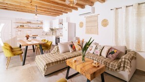 Un salon chaleureux avec un canapé, une table basse et des fauteuils jaunes. On aperçoit, au fond, une cuisine avec des meubles blancs et une table à manger.