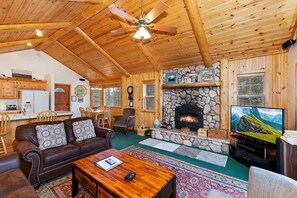 A log cabin interior with a stone fireplace, wooden furniture, and a television.