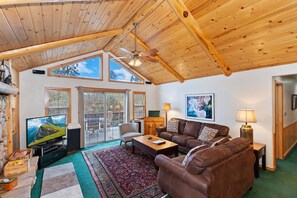A cozy living room with a wooden ceiling, large windows, a television, a brown leather sofa, and a coffee table.