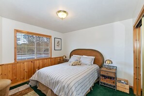 Un dormitorio con cabecera de madera, cama con ropa de cama a rayas, lámpara de noche, mesita de noche con lámpara, ventana con persianas e imagen enmarcada en la pared.