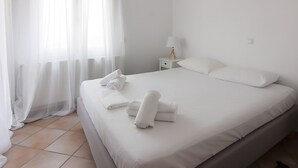 Een netjes opgemaakt bed met witte lakens en handdoeken, een witte nachtkastje met een lamp, en dunne gordijnen voor het raam.