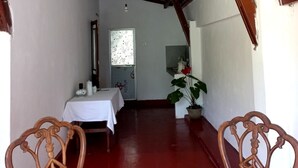 A room with a red floor, wooden ceiling beams, a white table with a white tablecloth, two wooden chairs, a potted plant, and a window with floral curtains. - Indra Sisila Villa Bentota  (Bentota)