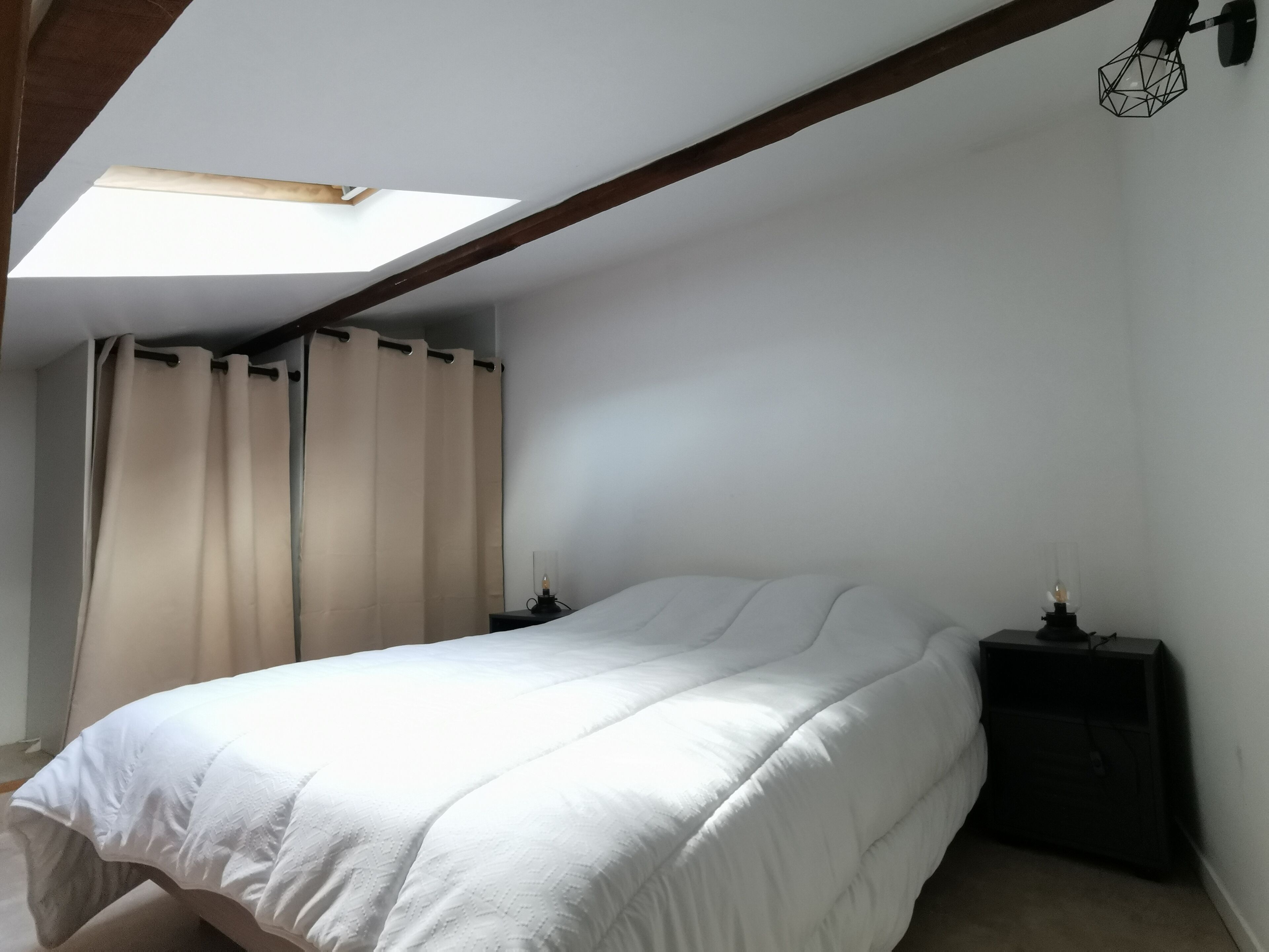 A bedroom with a skylight, a bed, two bedside tables, and a window with curtains.