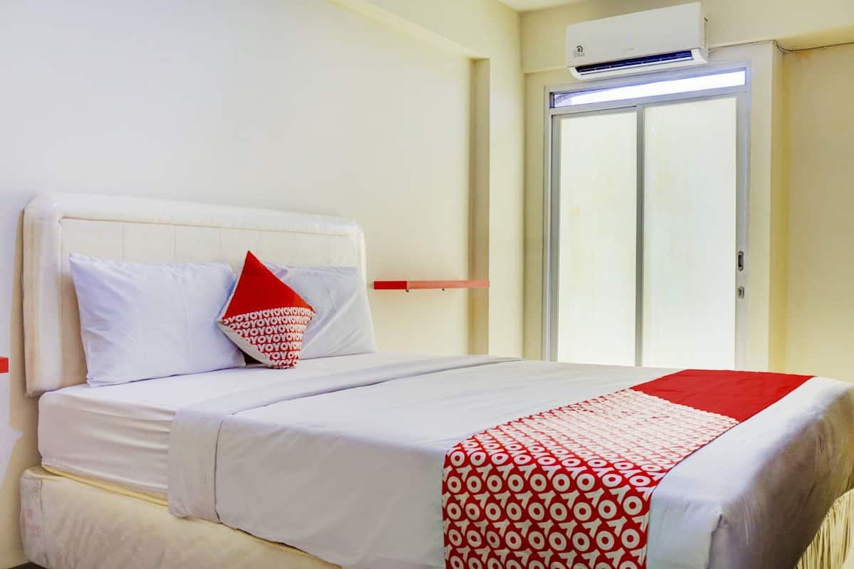 Habitación de hotel con una cama, un cojín de diseño rojo y blanco, una colcha blanca y roja, y una puerta con un panel de vidrio esmerilado.