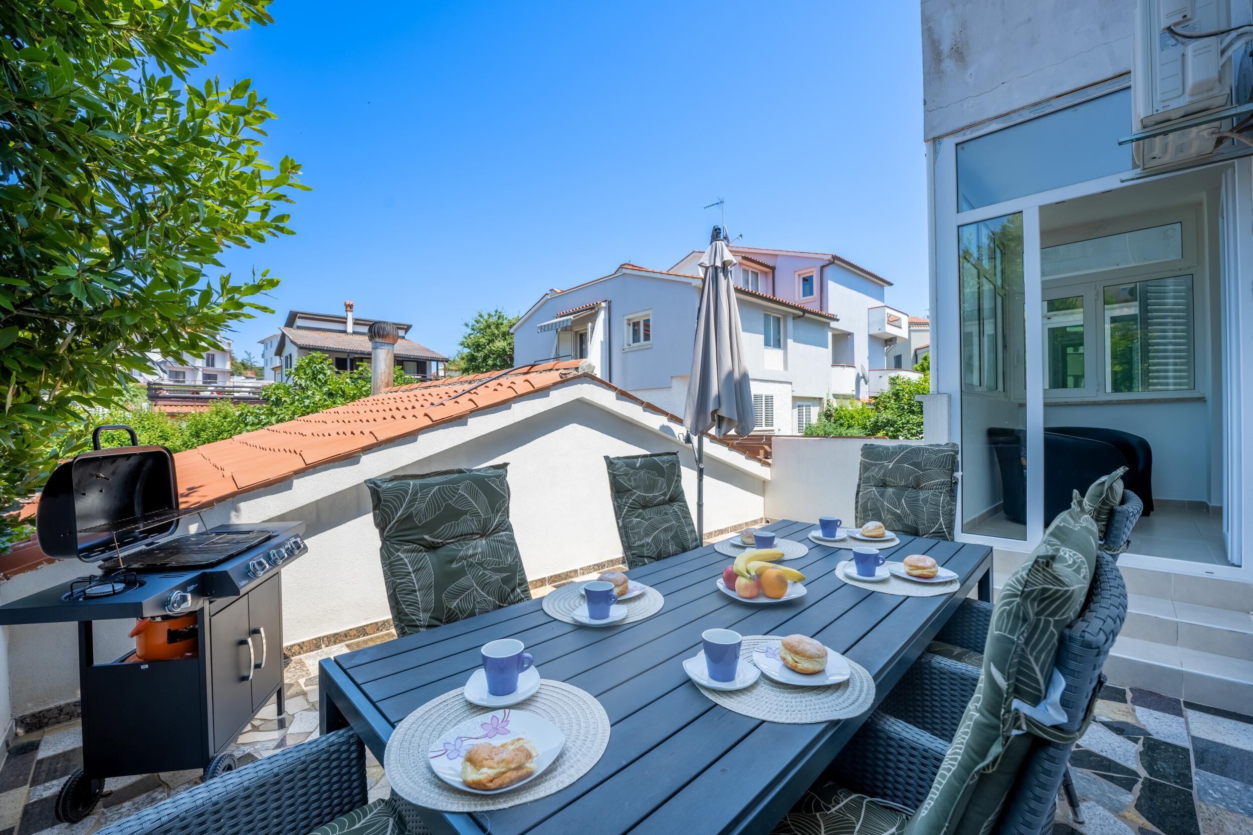 An outdoor dining area with a table set for two, a grill, and a view of neighboring buildings.