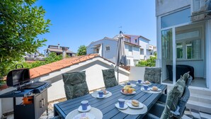An outdoor dining area with a table set for two, a grill, and a view of neighboring buildings.