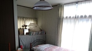 Ein Schlafzimmer mit zwei Betten, einem kleinen Tisch, einem Stuhl, einem Schreibtisch mit Stuhl, einer Pendelleuchte und einem Fenster mit Vorhängen.
