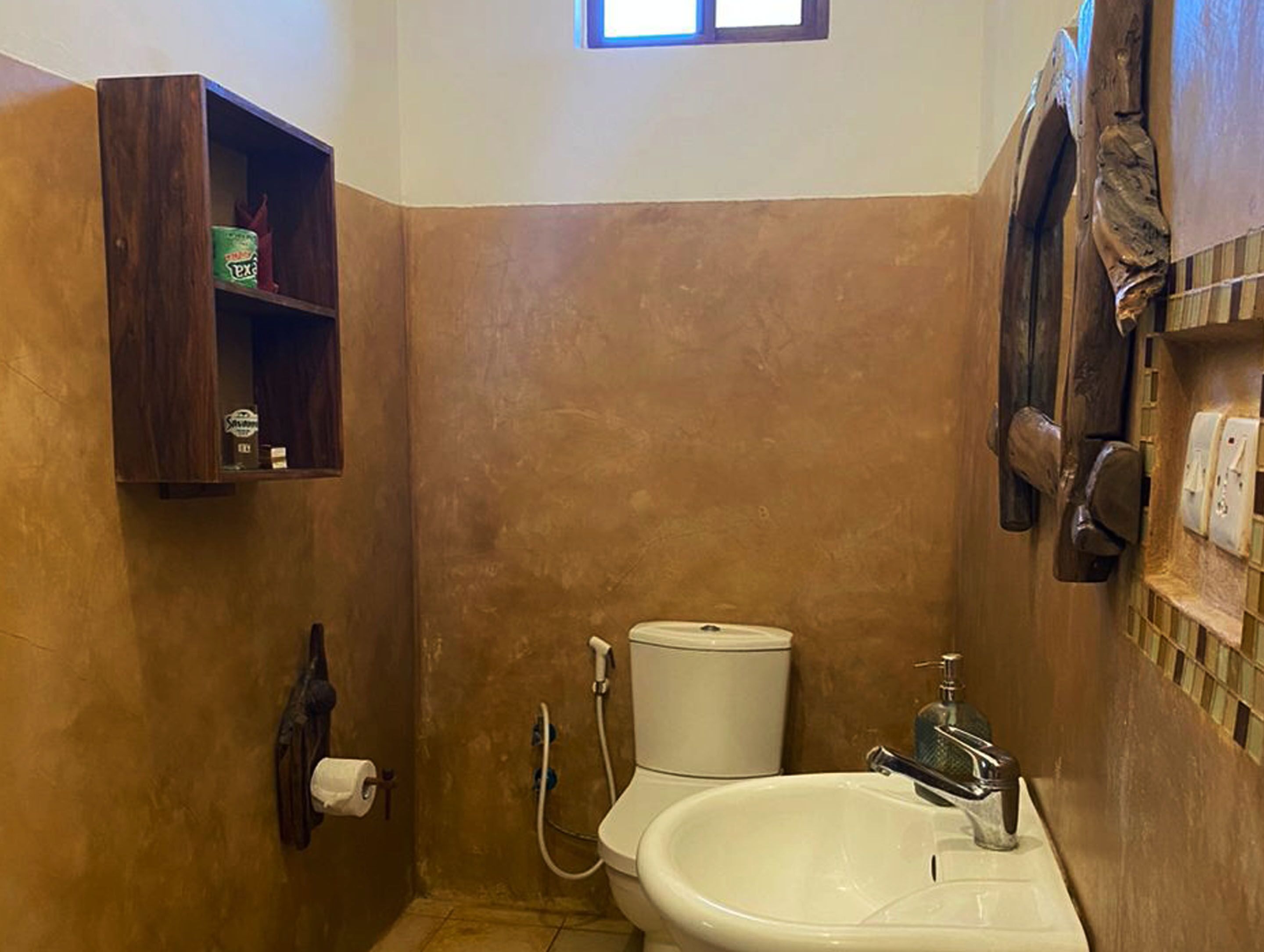 A small bathroom with a toilet, sink, and shower. There is a wooden shelf with a box of tissues and a bottle of hand soap.