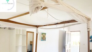A room with a bed under a canopy, a mirror, a small table, and a door leading outside. - Shamba Village Hotel (Paje)
