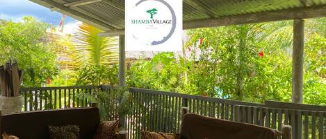 A covered outdoor seating area with a ShambaVillage sign, wooden furniture, and lush greenery in the background.