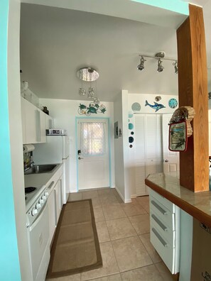 A kitchen with white cabinets, a stove, and a sink. There is a door with a blue frame and a window with a curtain. - Oceanfront Beach Luxury Retreat (St Thomas)