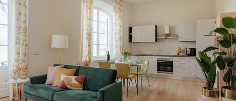A modern living room with a green sofa, a dining table with chairs, a kitchen area, and large windows with floral curtains.