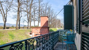 Ein Balkon mit schmiedeeisernem Geländer, ein Tisch mit Stühlen und Blick auf einen Park mit Bäumen und ein Gebäude in der Ferne.