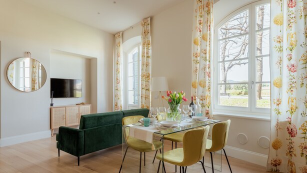 Ein Essbereich mit Glastisch, grünem Sofa und floralen Vorhängen.