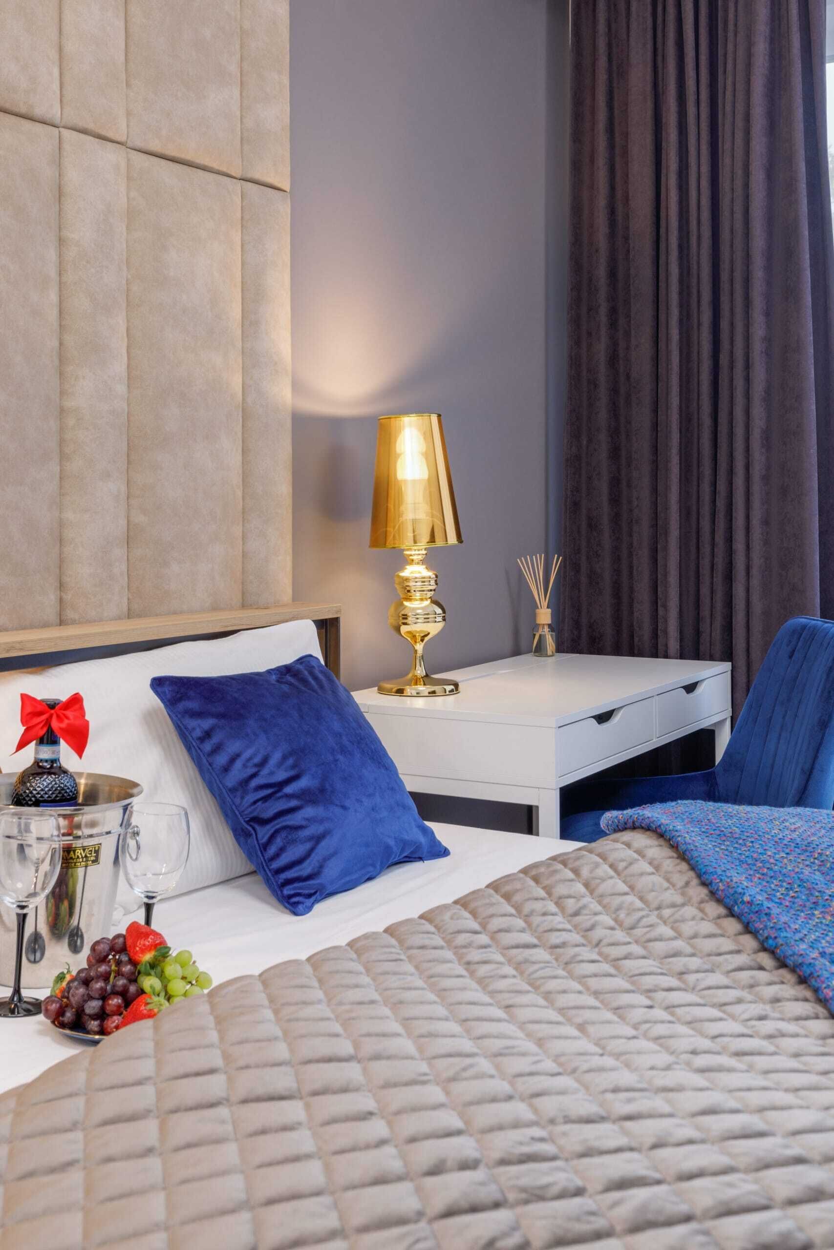 Hotellihuone, jossa on sänky, yöpöytä lampun kanssa, sininen tuoli ja pieni pöytä, jonka päällä on pullo ja lasi.