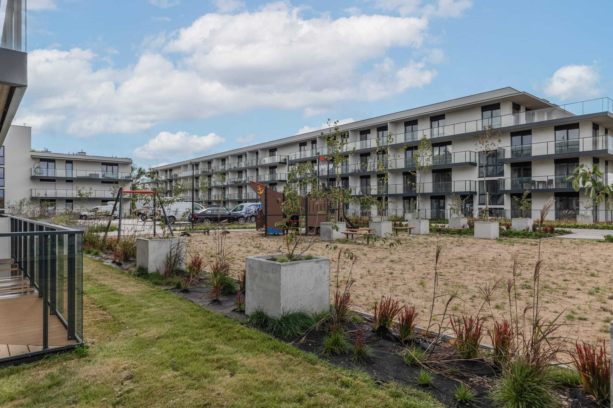 A modern multi-story building with balconies, a parking area, and landscaped garden beds.