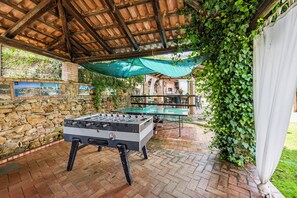 An outdoor area with a foosball table and a ping pong table under a wooden pergola, surrounded by stone walls and greenery.