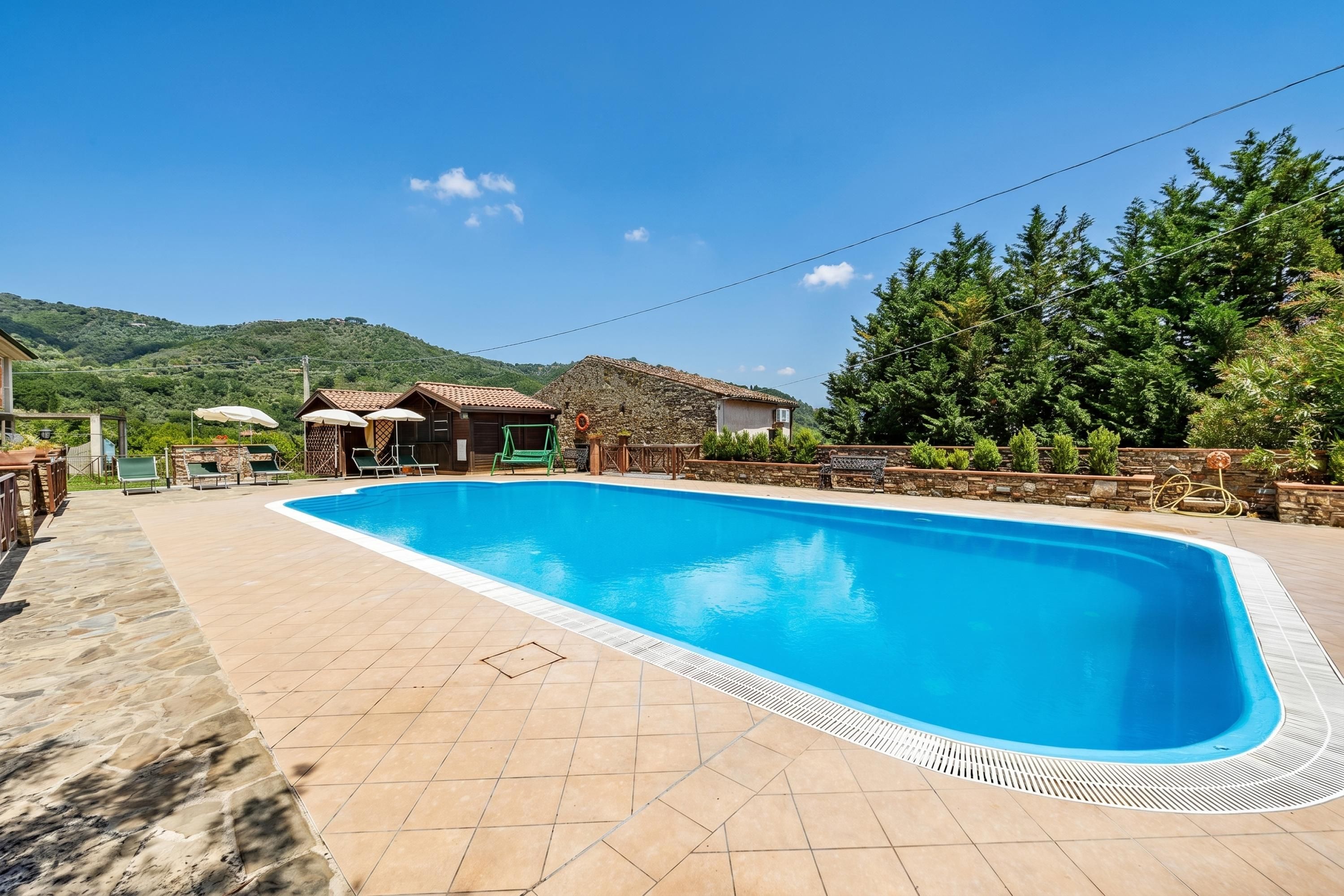 A swimming pool with a tiled deck, a stone building, and a view of a green hill.
