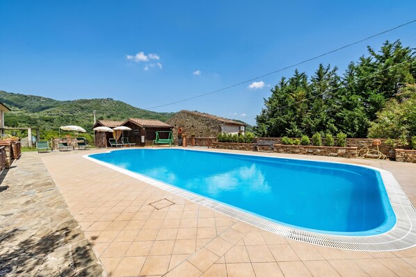 A swimming pool with a tiled deck, a stone building, and a view of a green hill.