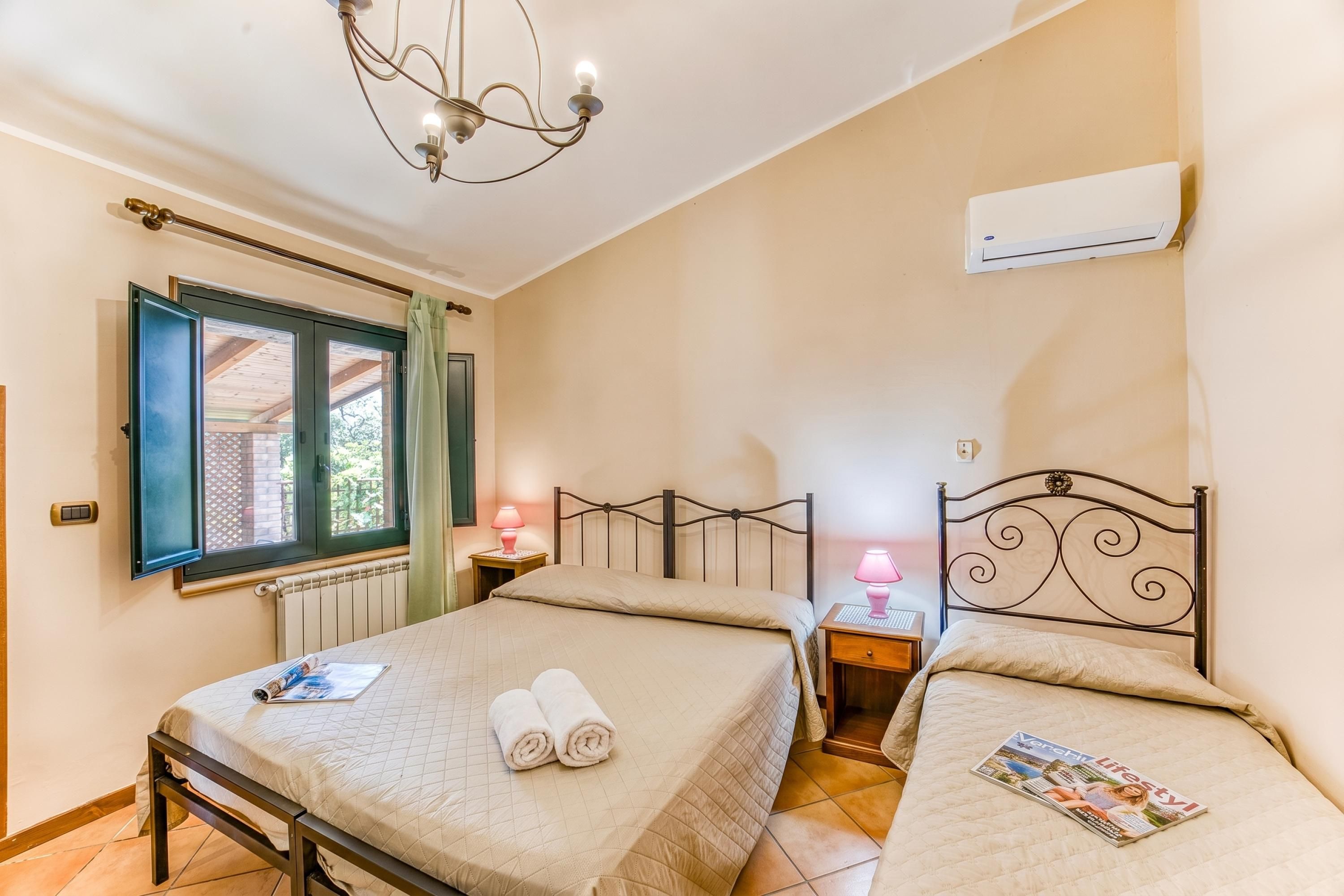 A bedroom with two beds, a chandelier, an air conditioner, and a window with curtains.