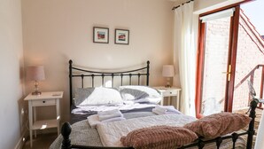 Un lit bien fait, recouvert d’une courtepointe à carreaux, avec deux oreillers et un plaid rose. On y trouve deux tables de chevet avec des lampes, un petit tiroir et une fenêtre avec des rideaux.
