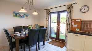 Un coin repas avec une table dressée pour deux personnes, une horloge au mur et une vue sur le jardin par la porte.