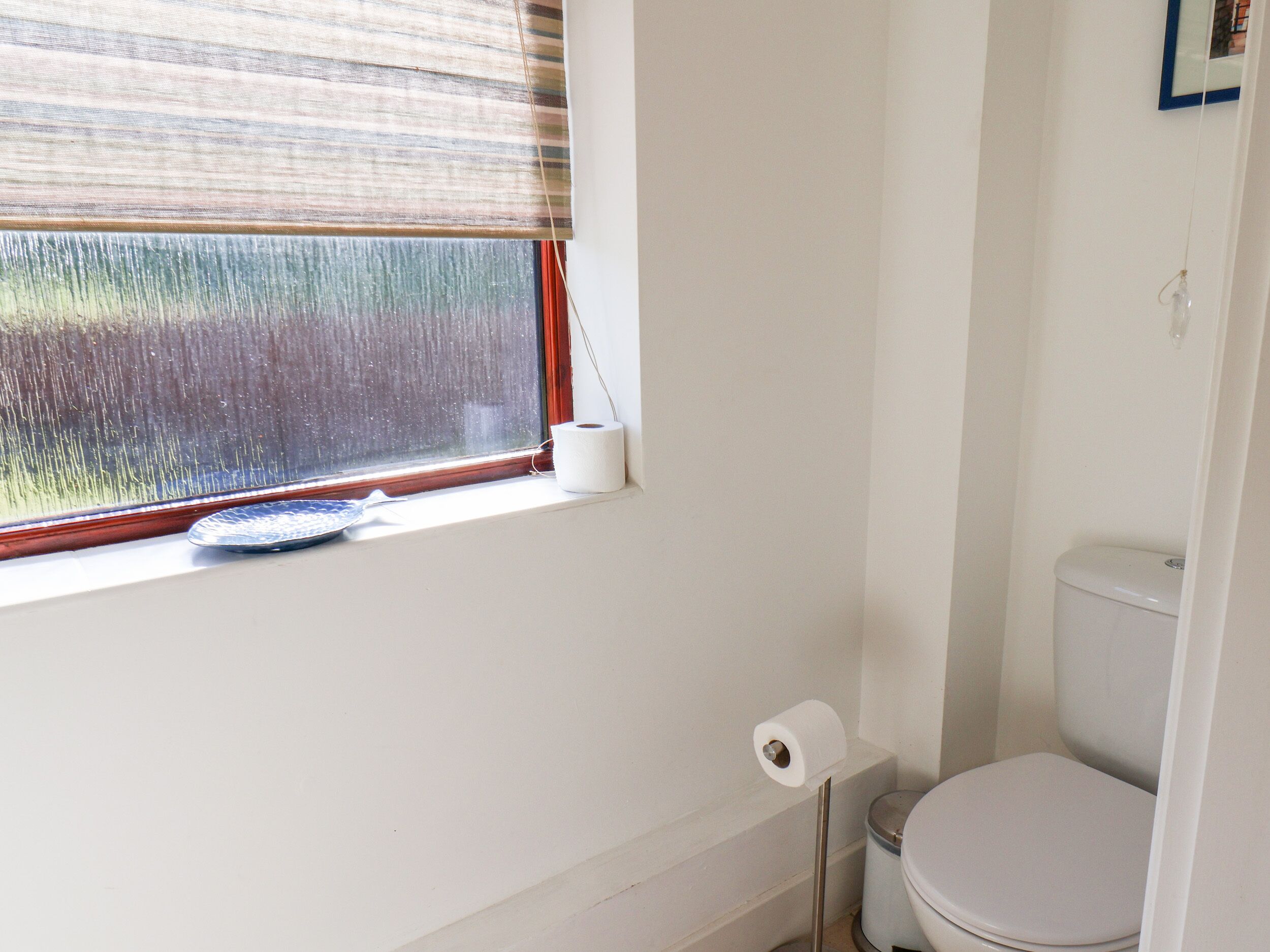 Une salle de bain avec des toilettes, une fenêtre avec un store et un rouleau de papier toilette par terre.
