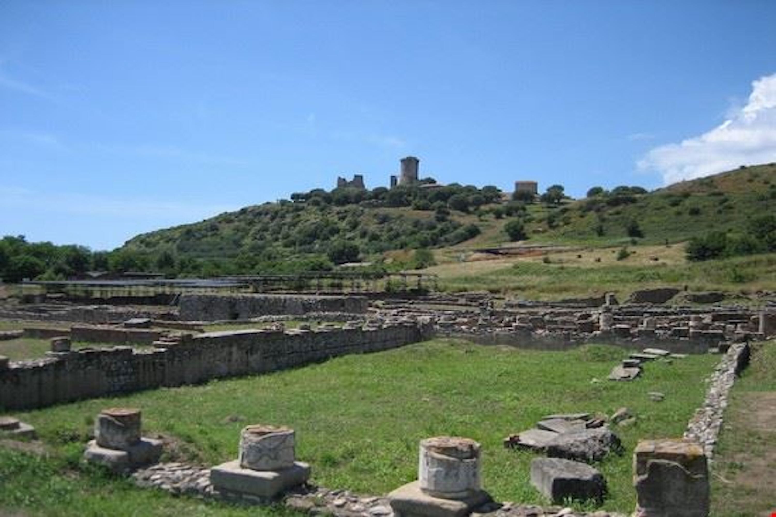 Ancient ruins with stone structures and walls, a hill with a tower, and a grassy area.