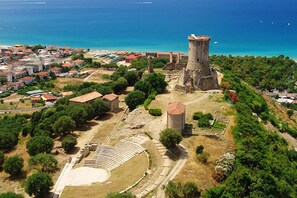 An ancient amphitheater with ruins and a tower, surrounded by trees and a coastal town.