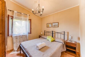 A bedroom with a bed, a nightstand, a lamp, a chandelier, a window with curtains, and two wall-mounted pictures.