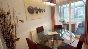 Un espace repas moderne avec une table en verre, des chaises, un vase contenant des fleurs séchées et une horloge murale.