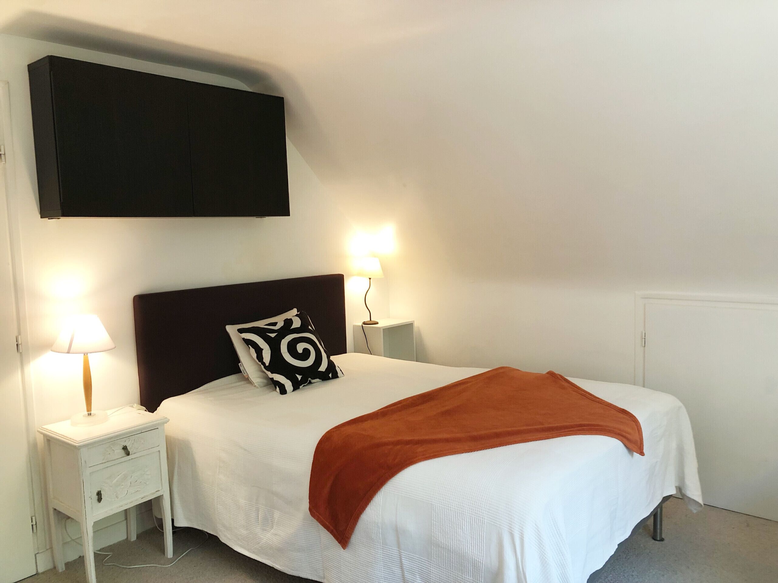 Ein ordentlich bezogenes Bett mit weißer Decke und einer orangefarbenen Tagesdecke, begleitet von weißen Nachttischen mit Lampen.