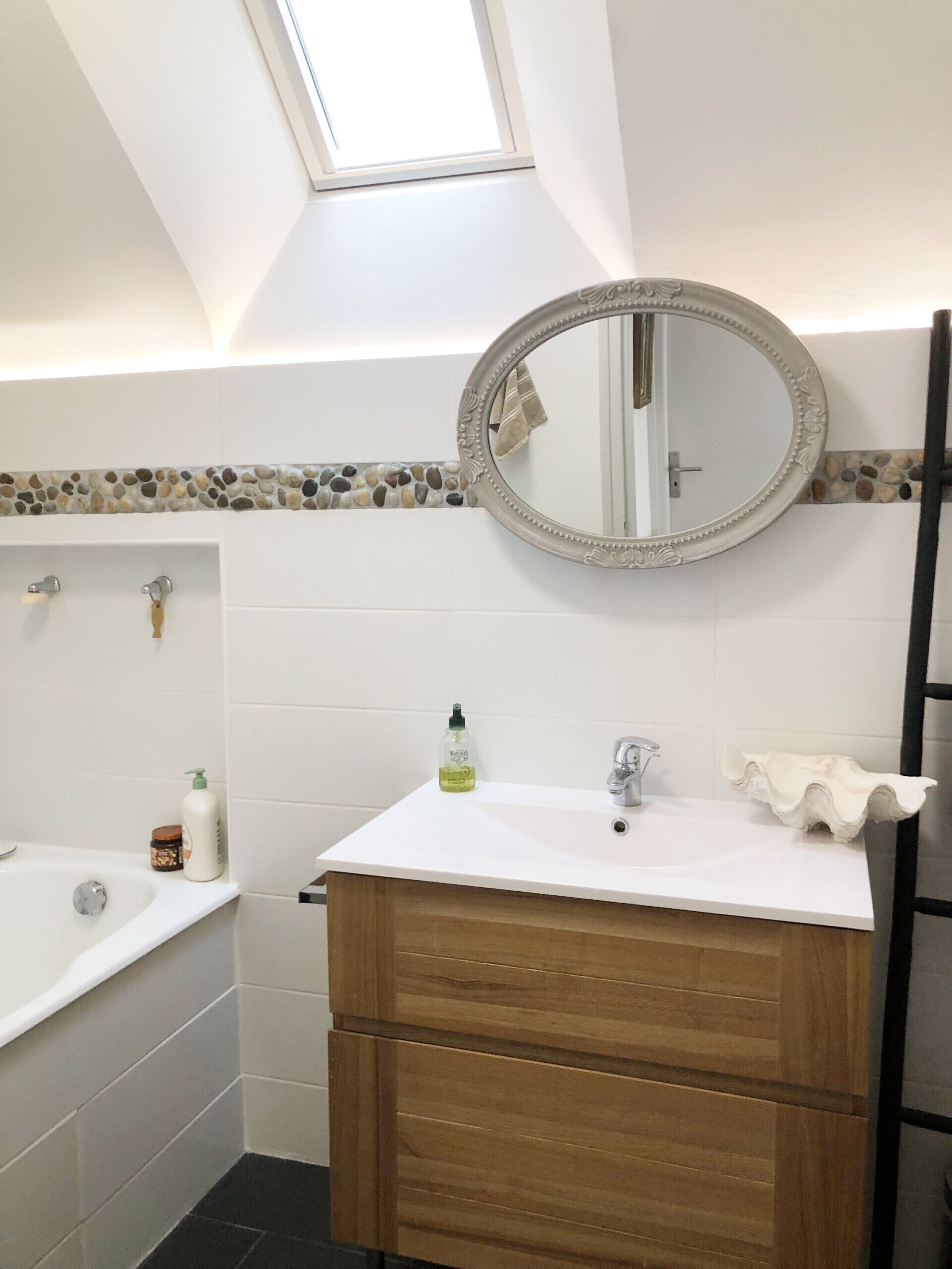A bathroom with a skylight, a round mirror, a white sink, wooden vanity, and a bathtub.