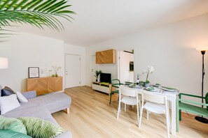 A modern living room with a sofa, dining table, and chairs, a kitchen area with cabinets, and a television.