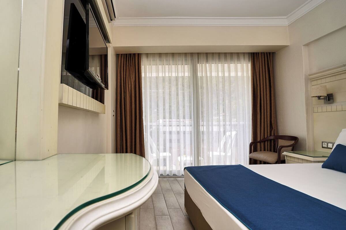A hotel room with a bed, a glass table, a chair, a TV, and a window with curtains.