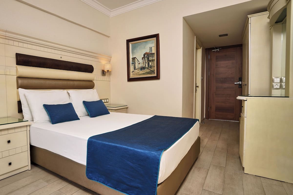 A hotel room with a bed, bedside tables, a nightstand, a desk, a chair, a painting on the wall, and a door leading to a kitchen.