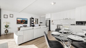 A modern living room with a white sofa, a dining table set for two, and a kitchen area with white cabinets and stainless steel appliances. - Edge One - Dog Friendly (Jindabyne)