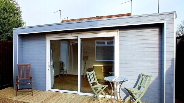 Ein modernes Gebäude mit einer Schiebetür, die zu einem Innenraum führt, einer kleinen Terrasse mit Tisch und zwei Stühlen sowie einem Holzdeck.