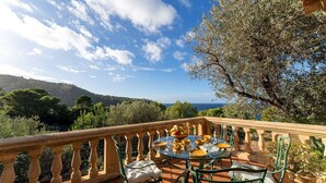 A terrace with a round glass table set for dining, surrounded by green chairs, overlooking a scenic landscape with trees and a view of the sea. - Casa Perdita, ideal for family and relaxed vacations (Deiá)