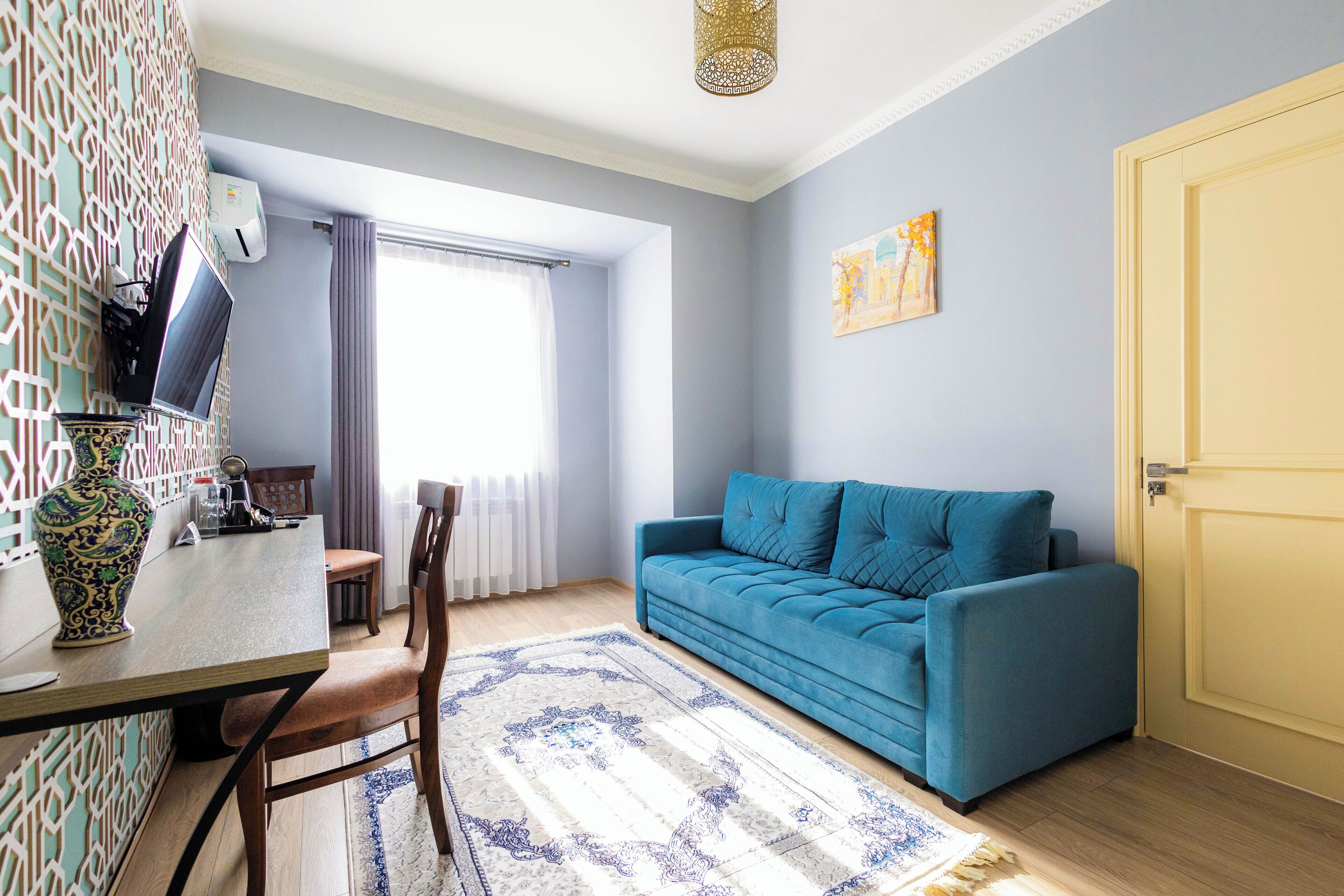 Una sala de estar con un sofá azul, una mesa de comedor con sillas, un tapete estampado y un jarrón decorativo.
