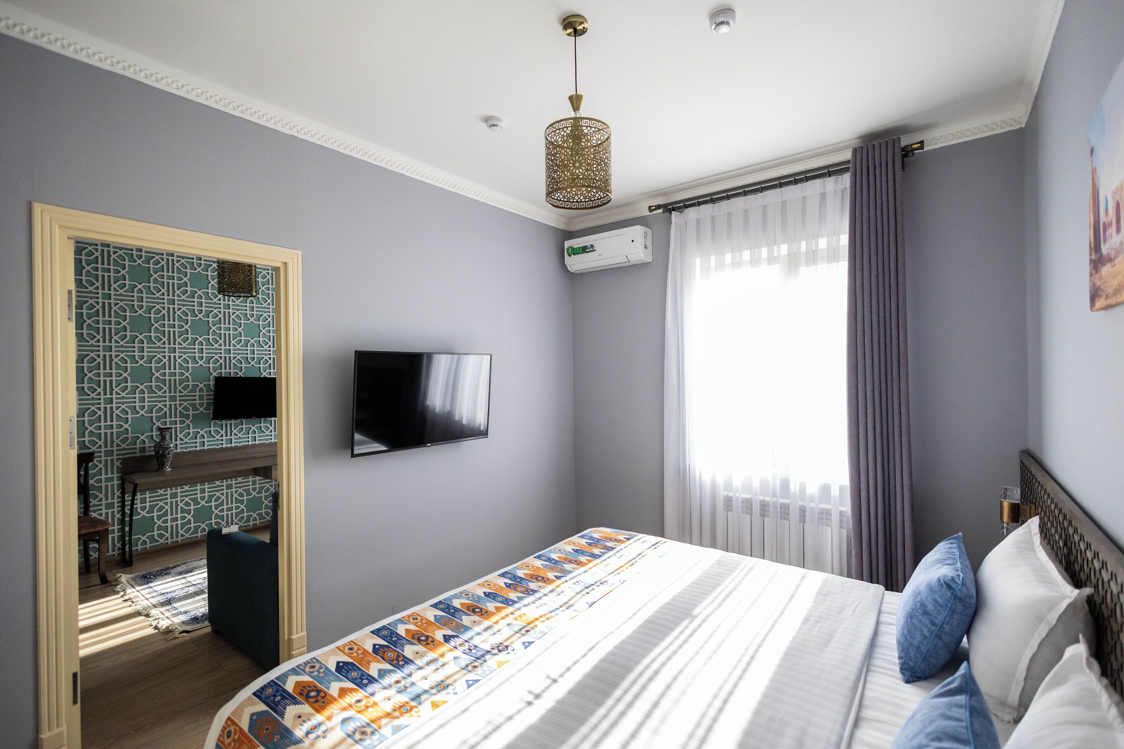 A bedroom with a bed, a TV mounted on the wall, a window with curtains, and an air conditioning unit.