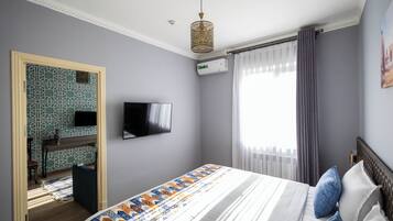 A bedroom with a bed, a TV mounted on the wall, a window with curtains, and an air conditioning unit.