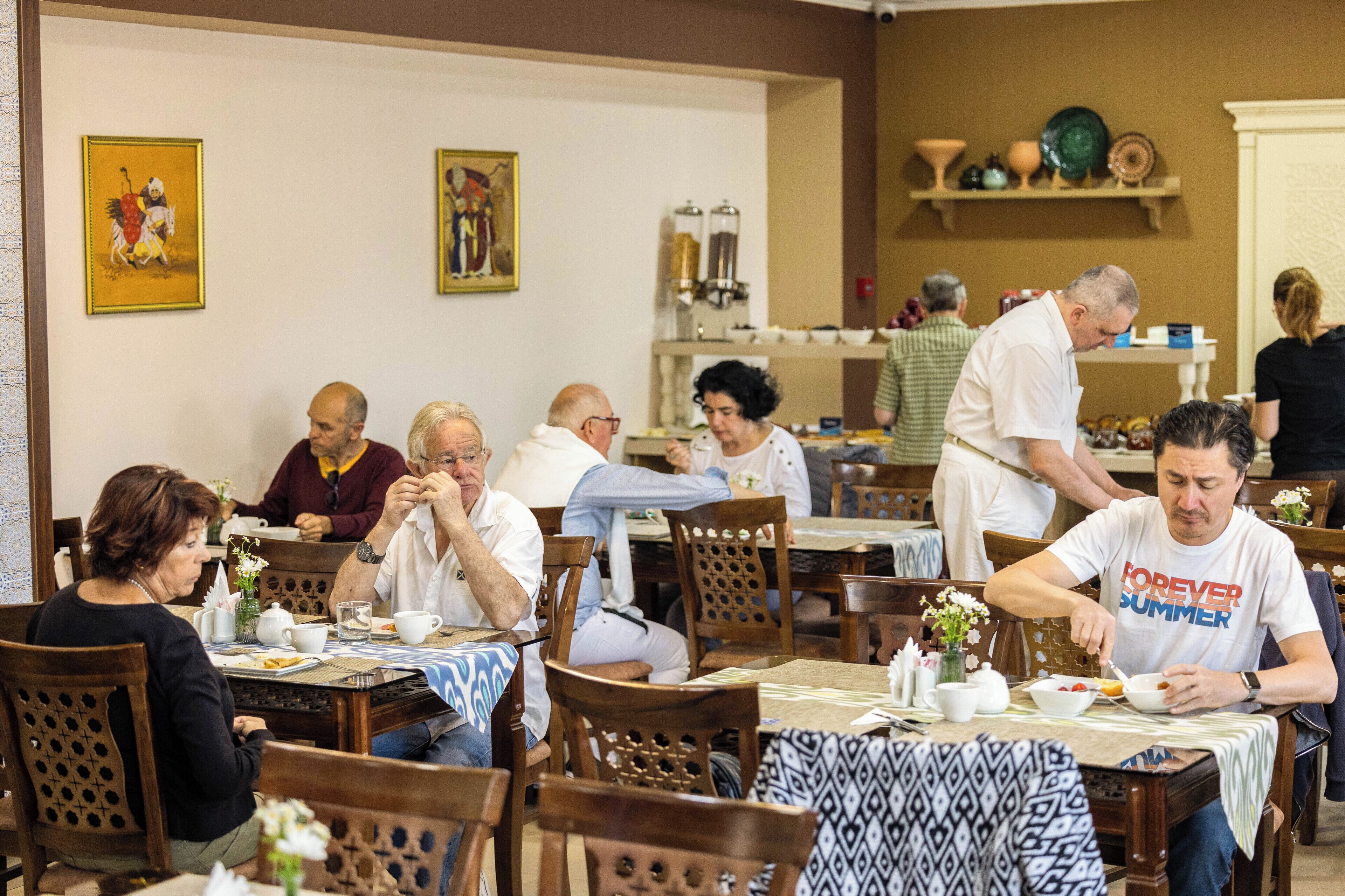 A dining area with wooden tables and chairs, a waiter serving food, and decorative items on shelves.