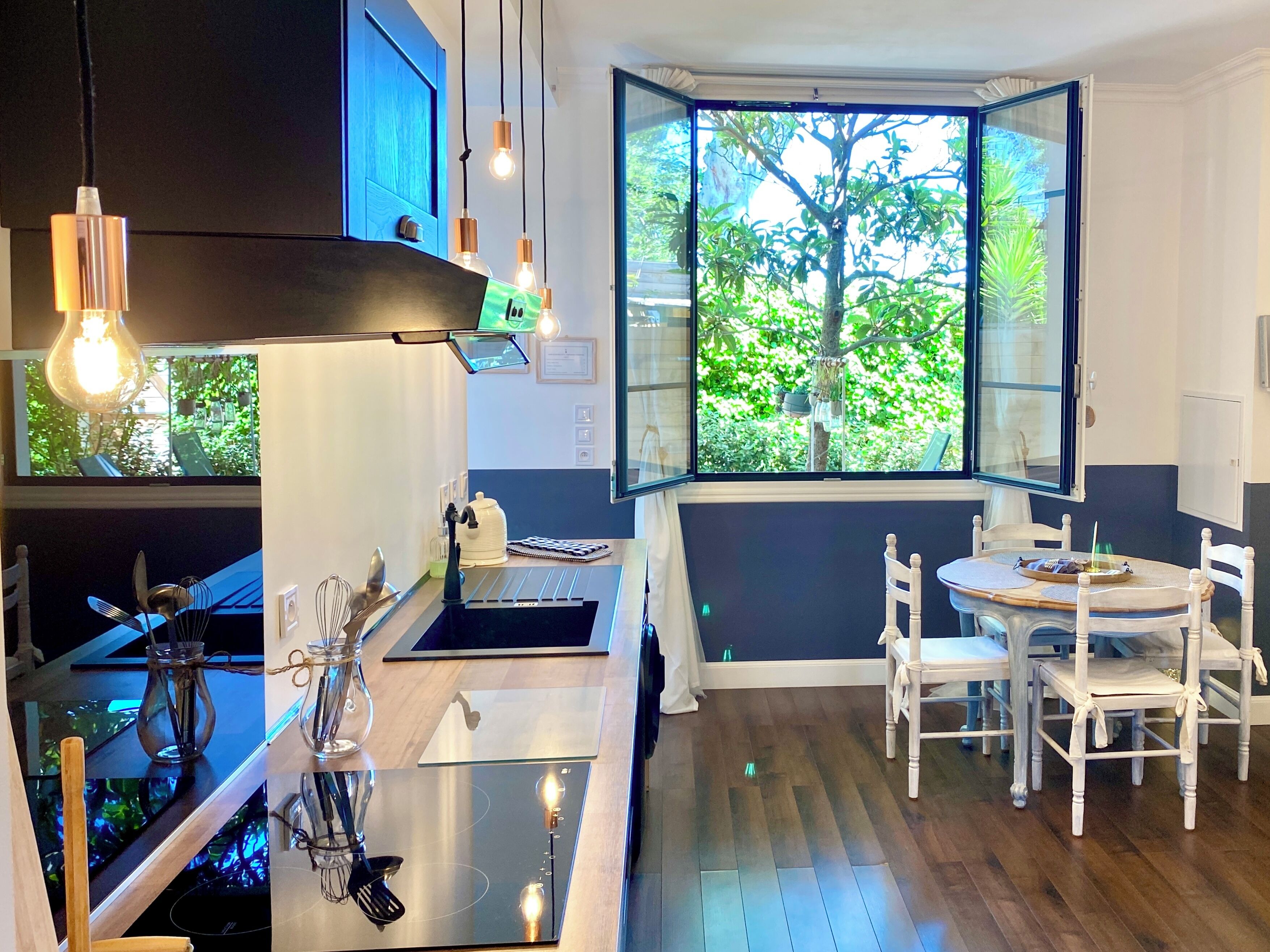 Een moderne keuken met een centraal eiland, een eethoek met witte stoelen, en een groot raam met uitzicht op groen.