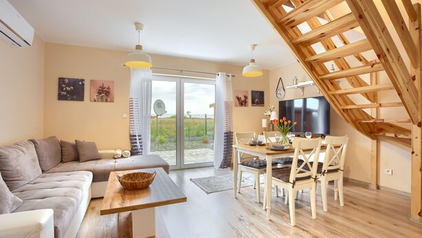 Een moderne woonkamer met een bank, eettafel en stoelen. Er is een grote televisie en een houten plafondconstructie.