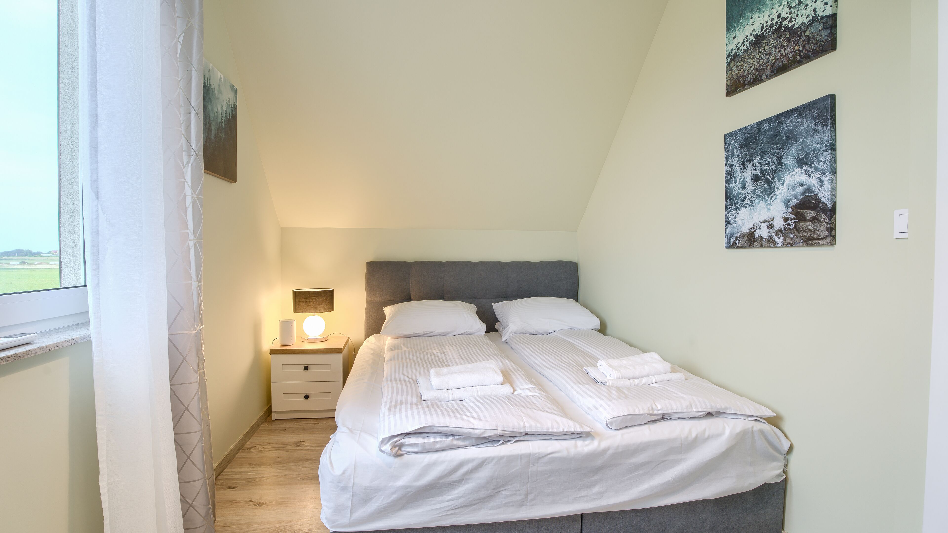 Una cama individual bien hecha con sábanas blancas y dos almohadas, una mesita de noche con una lámpara y dos cuadros en la pared.